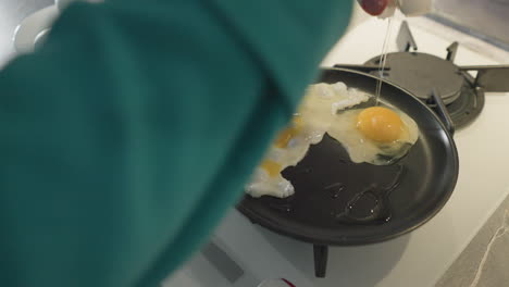 older woman cracking egg into frying pan in modern kitchen, preparing meal with fresh ingredients, visible eggs on plate, kitchen tools, and stove with focused cooking action