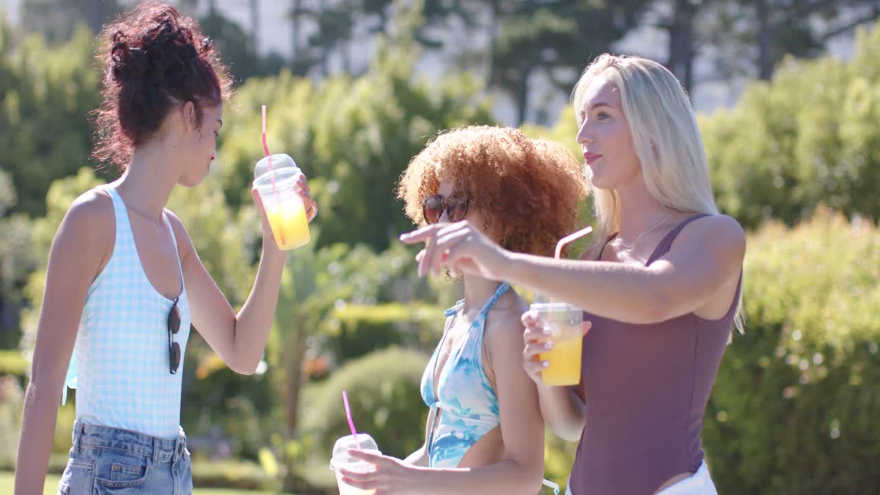 Female friends in 20s arriving poolside, holding plastic cups, adjusting sunglasses and celebrating