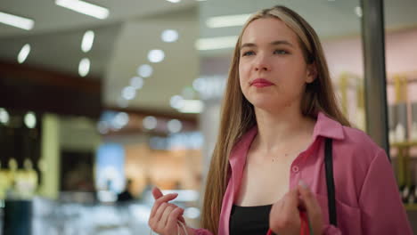 hermosa dama en vestido rosa levanta sus bolsas de compras rojas y verdes menta mientras toma una respiración profunda con una cálida sonrisa en su cara mientras camina a través de un centro comercial bien iluminado, el fondo está borroso