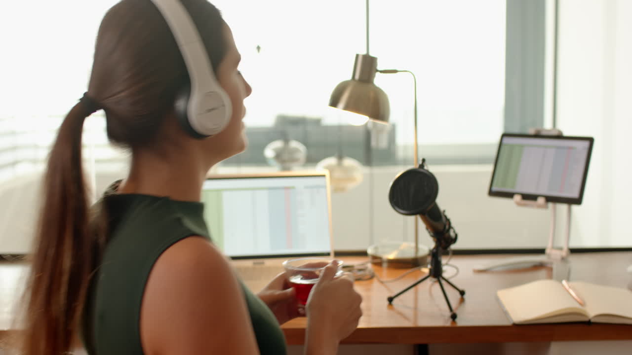 Wearing headphones, woman holding drink and working on laptop at home office