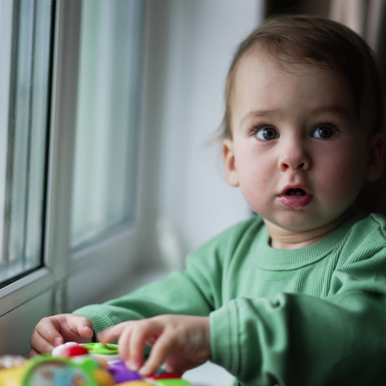 Lovely child standing at the window-sill with a toy in front of him. Cute baby pressing the buttons on the toy and then looks at camera. Close up