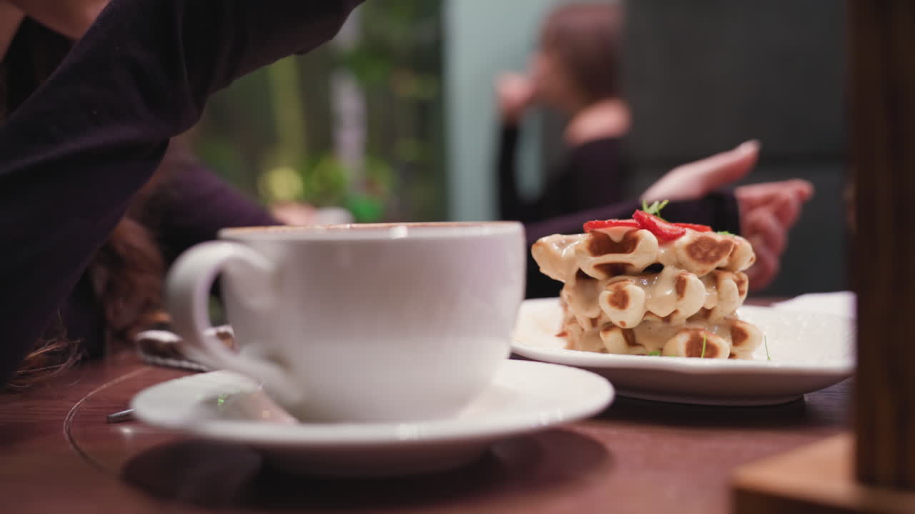 Lady enjoys waffles using fork at cozy cafeteria table with stacked dessert topped with strawberries and creamy sauce, alongside white cup and saucer, capturing relaxed moment of indulgence