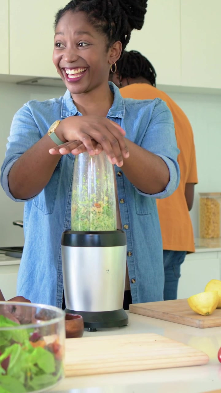 Video verticale: Donna in jeans che blocca la tazza del frullatore in cucina, frullando verdure per l'insalata.