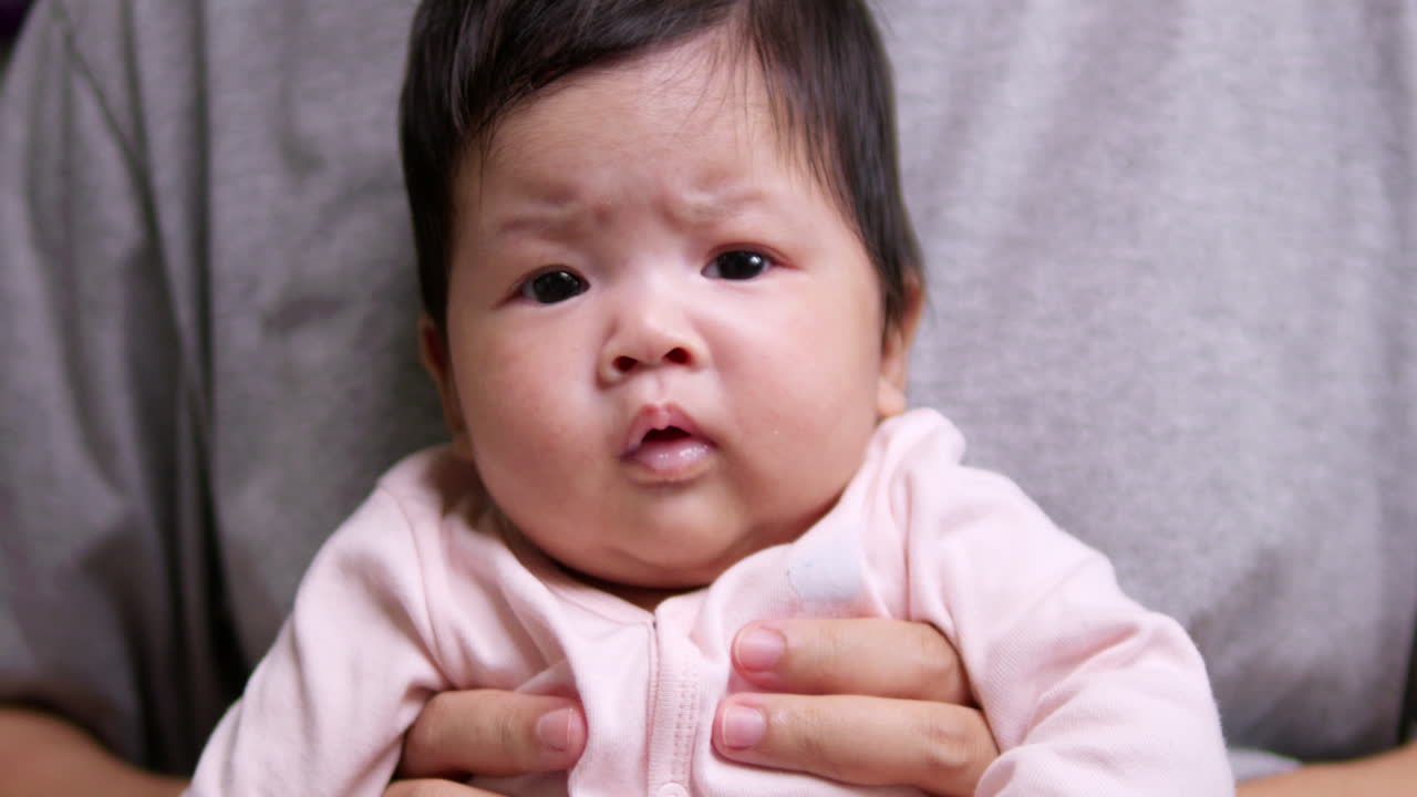 A newborn baby is furrowing her brows and squirming while her mother is holding her down on her lap.