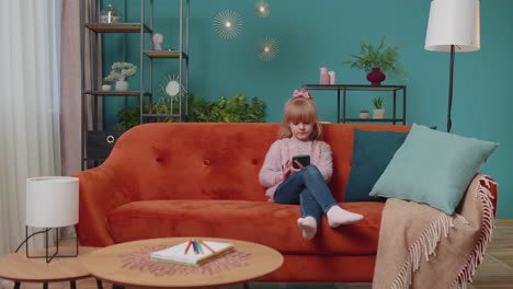 little elementary school kid girl using mobile phone technology device sitting on sofa alone at home