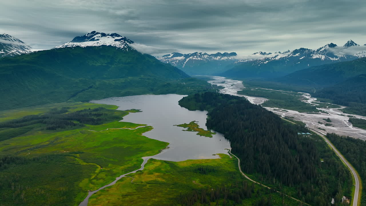 Approaching the river in the beautiful green valley. Snow-capped mountains covered with clouds at backdrop. Gloomy day in Alaska, USA. Aerial view