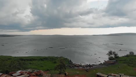 Toma-Aérea-De-Cabañas-De-Aldea-A-Orillas-Del-Lago-Victoria-Con-Nubes-De-Tormenta-Que-Se-Extienden-Hacia-El-Lago