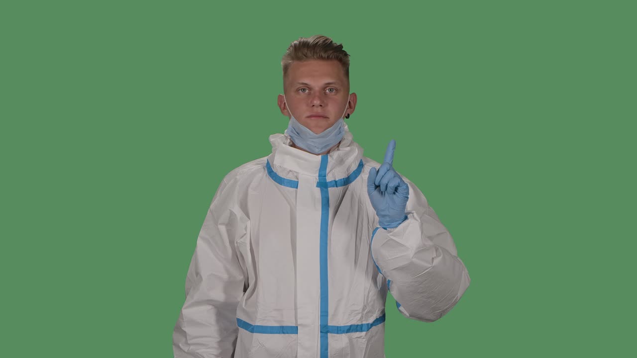 Portrait of a man in a protective suit and medical mask looks into the camera and shows how to properly wear a medical mask. Green screen, chroma key. Slow motion. Close up