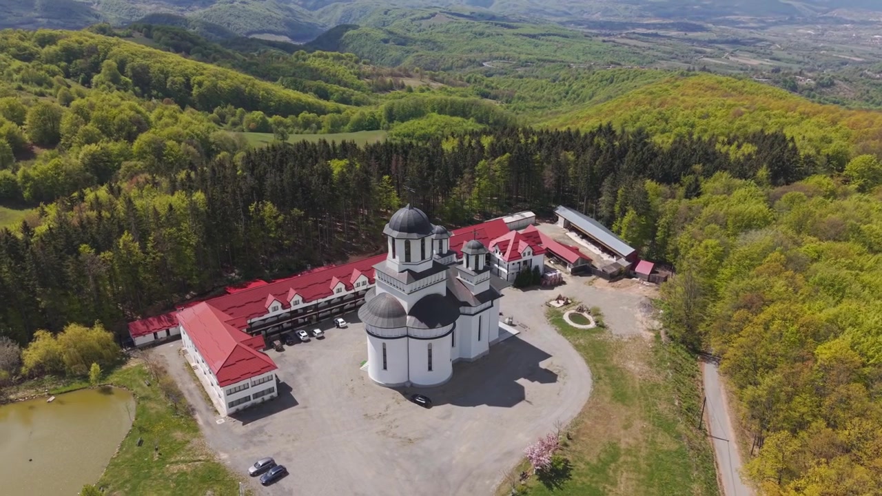 Aerial view of a monastery complex centered around a white church with dark domes, surrounded by lush green forests and rolling mountains.