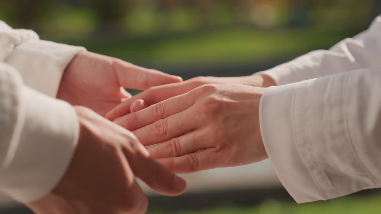 屋外で結婚指輪を交換する高齢のカップル。柔らかな日差しが二人の優しい手を温めています。庭園を舞台にした誓いの言葉の交換がクローズアップされ、生涯の誓いと愛情を伝える優しい触れ合いが親密な雰囲気を醸し出しています。