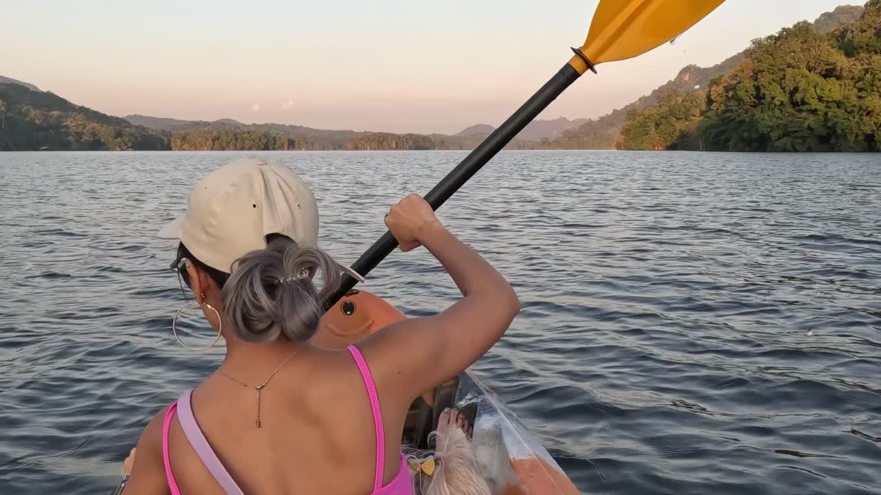 A person paddles a kayak on a calm lake, surrounded by lush green hills under a clear sky.