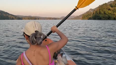 a person paddles a kayak on a calm lake, surrounded by lush green hills under a clear sky.