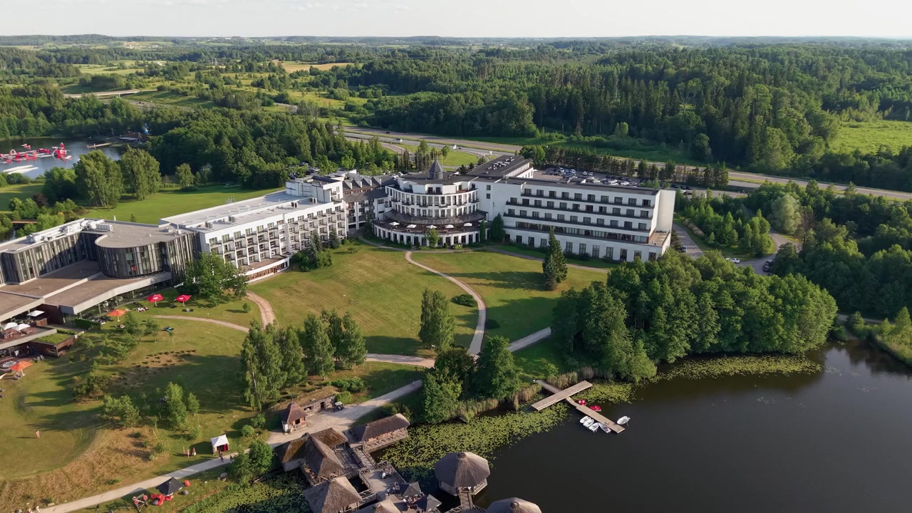 Drohnenvideo des Vilnius Grand Resort bei Vilnius, Litauen, zeigt moderne Architektur, idyllische Seelage, üppiges Grün und wunderschöne sonnige Landschaften mit weitläufiger Weitwinkelkomposition.