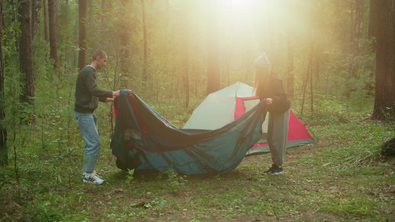 Young couple unfolds tent fabric on forest ground during camping setup as soft sunlight streams through tall trees in background, creating warm and serene outdoor
