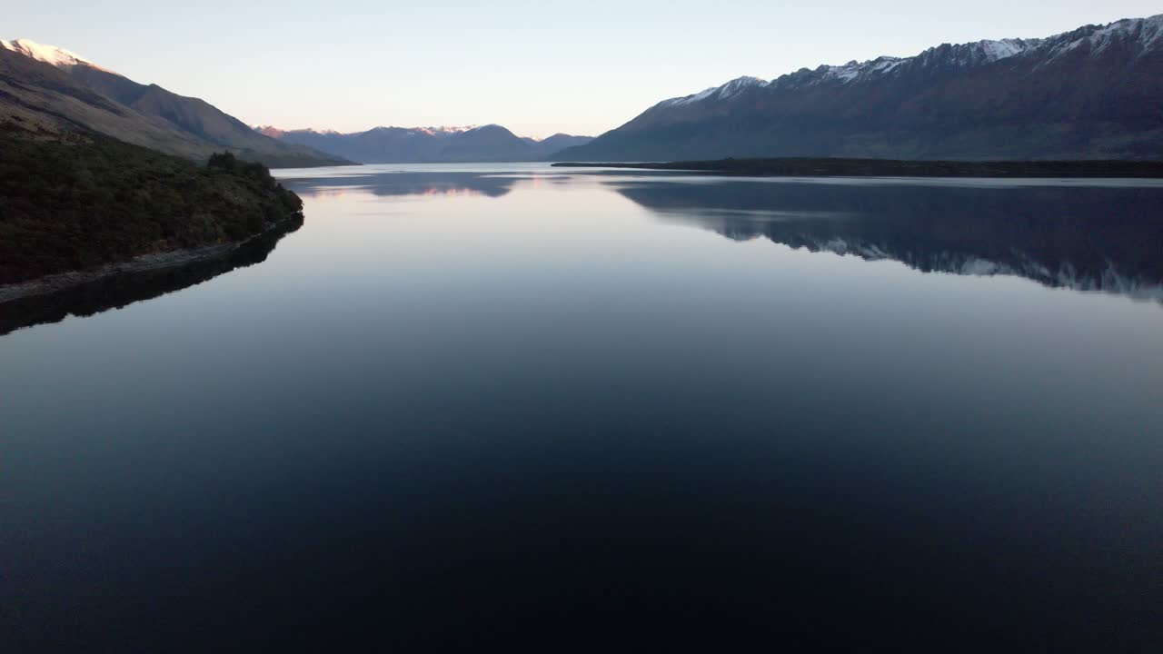 Aerial flying over Lake Wakatipu with the lake surrounded by snow capped mountains reflecting in the lake just after sunset in Queenstown New Zealand