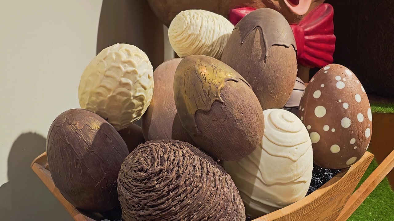 Assorted chocolate Easter eggs with detailed patterns and textures in a wooden basket, displayed beside a bunny decoration in warm indoor light for seasonal candy shop video and spring decor