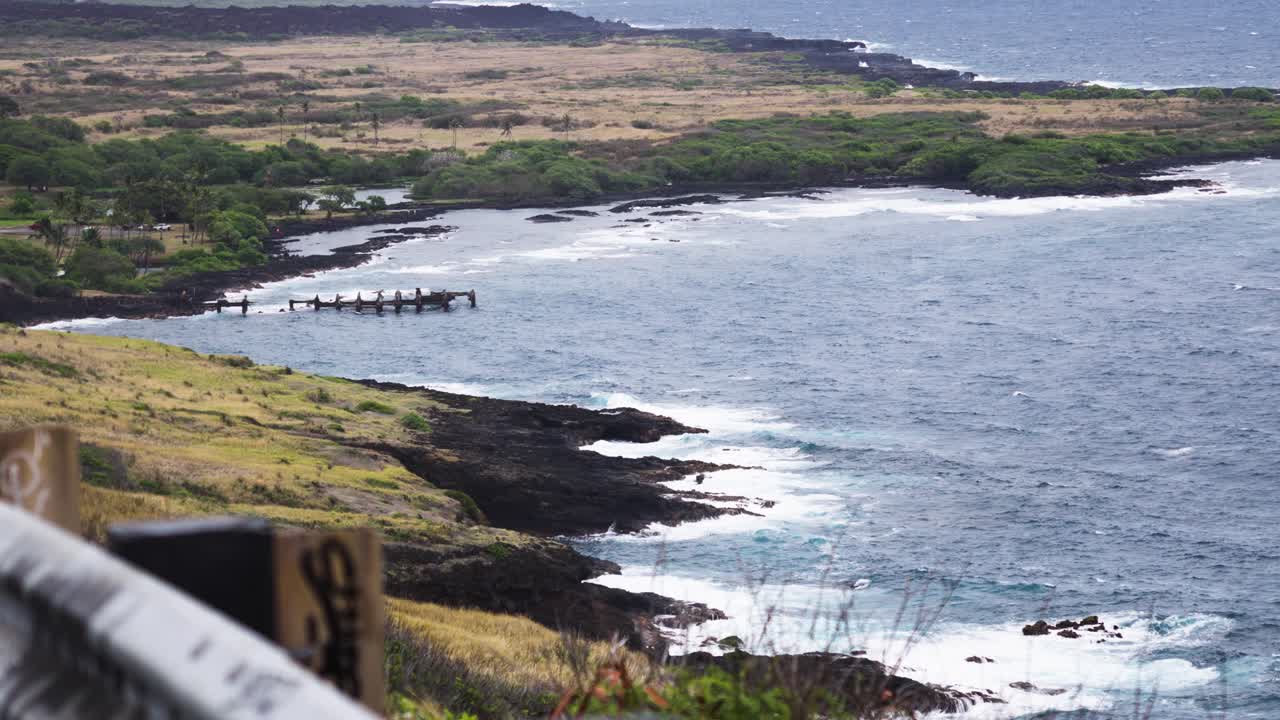 From a clifftop lookout, gentle swells trace a lava-fringed shoreline where weathered pier pilings jut from a quiet cove, with scrubby plains and green thickets under soft, hazy daylight