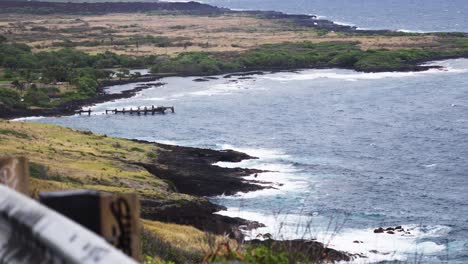 from a clifftop lookout, gentle swells trace a lava-fringed shoreline where weathered pier pilings jut from a quiet cove, with scrubby plains and green thickets under soft, hazy daylight
