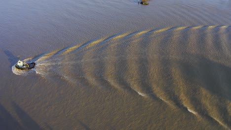 Aerial-birds-eye-view-tug-boat-passing-by-a-commercial-river-causing-waves-on-a-twilight-sunset-muddy-water-reflections-in-the-water-with-shadows-on-a-commercial-straight-boating-on-the-job-on-route