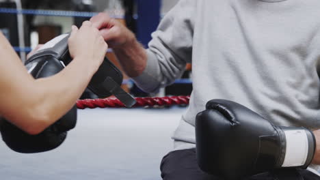 female personal trainer helping senior male boxer putting boxing gloves on in gym