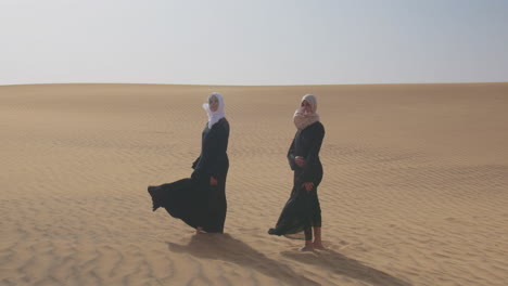 Dos-Mujeres-Musulmanas-Con-Vestido-Tradicional-Y-Hiyab-De-Pie-En-Un-Desierto-Ventoso-Y-Mirando-A-La-Cámara