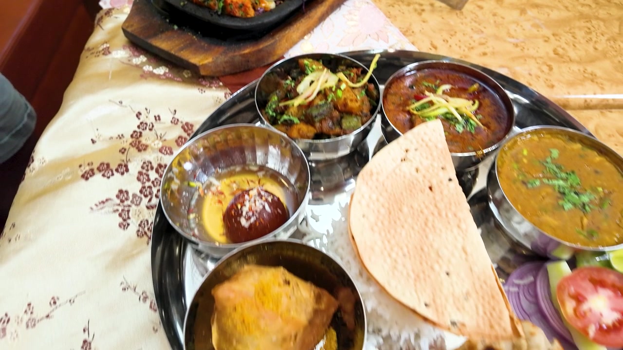 High-angle pan over sizzling seekh kebabs, thali platters, and samosas on a restaurant table.