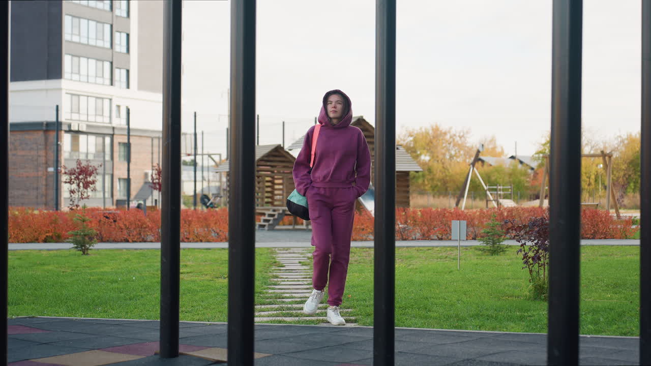 View between iron bars of sportswoman carrying bag walking towards building with excited expression on face in playground setting during autumn lighting with shadows and foliage backdrop