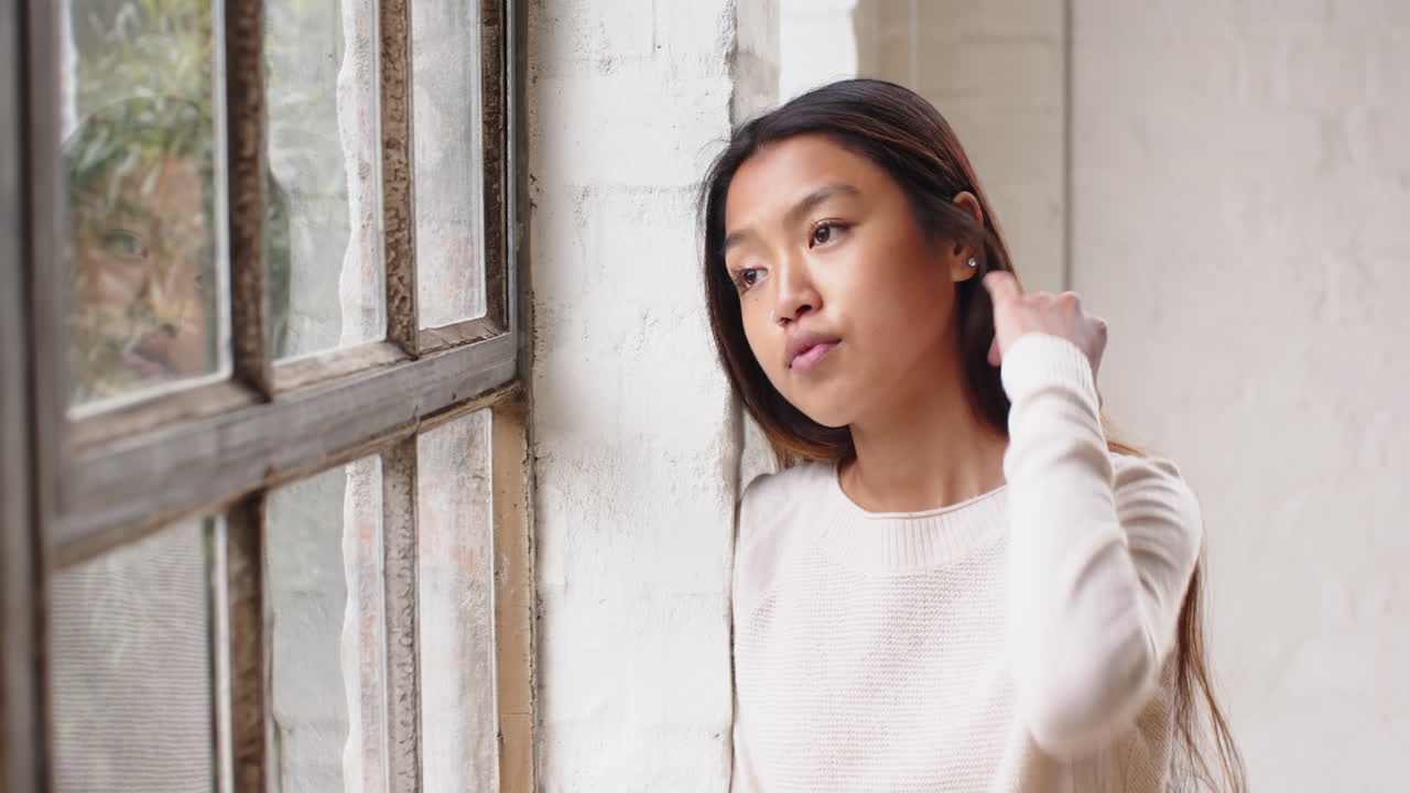 Asian young woman gazing out window, reflecting thoughtfully at home