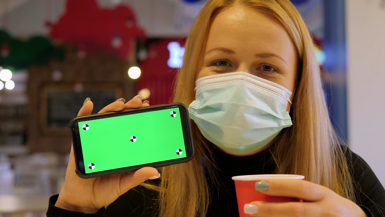 A woman in a medical mask holds a phone with a chroma key in a cafe.