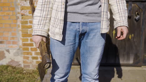 close-up view of a man in jeans holding an ax outside a country house