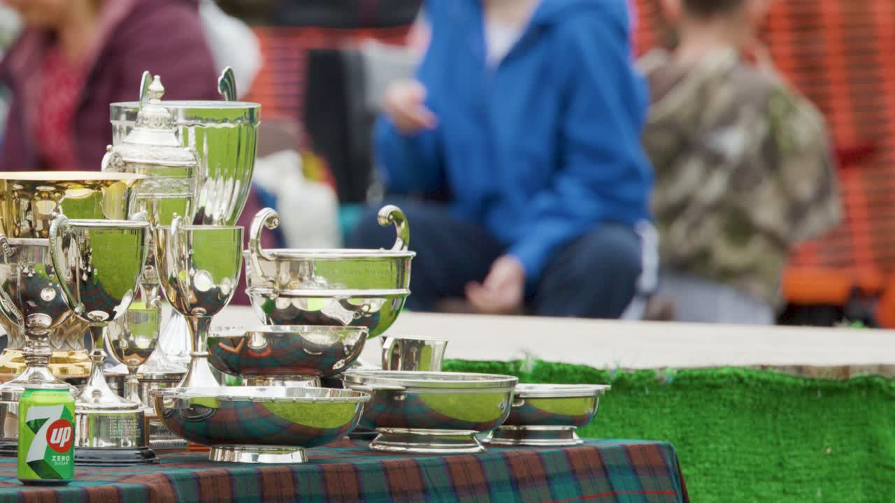 Silver trophies and awards displayed on a tartan-covered table outdoors, with people in kilts and casual clothing in the background, soft daylight, static camera
