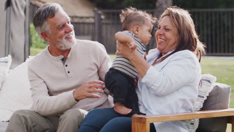 Senior-Hispanic-couple-sitting-on-a-seat-in-the-garden-passing-their-baby-grandson-over,-close-up