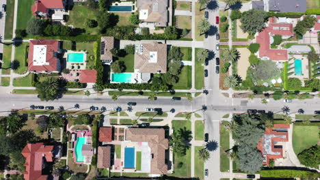 drone shot flying over beautiful palm tree lined street following black car driving, the street is surrounded by mansions with swimming pools in west hollywood, los angeles, california