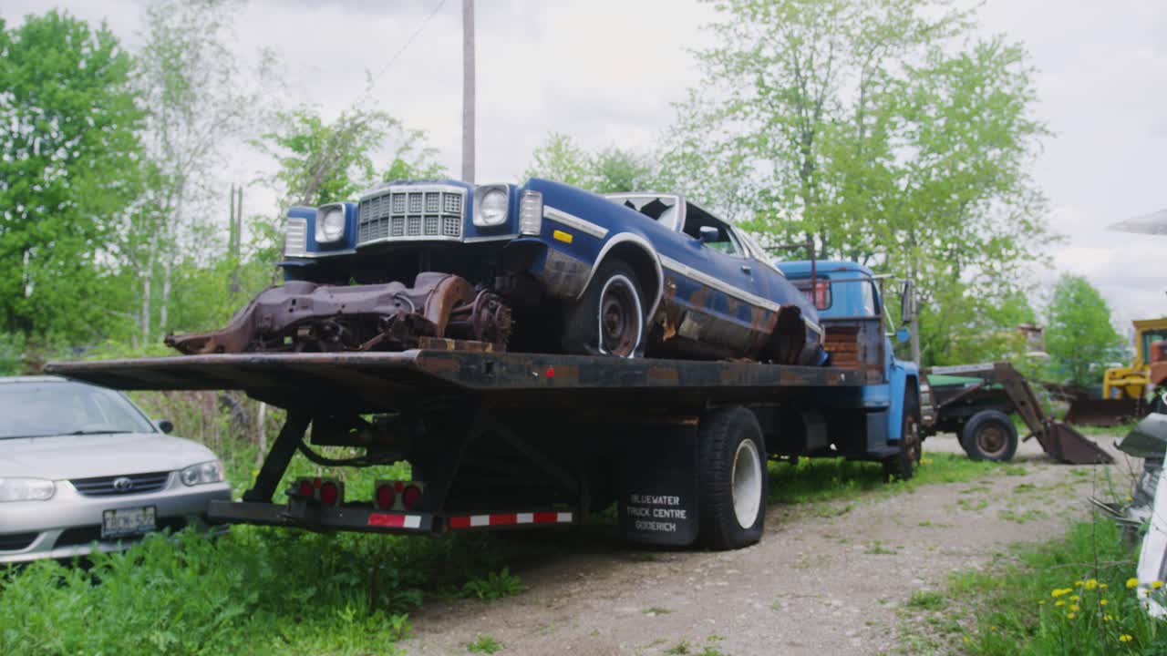 форд торино 1970-х, сидящий на задней части флатона, окруженный скрап-автомобилями.