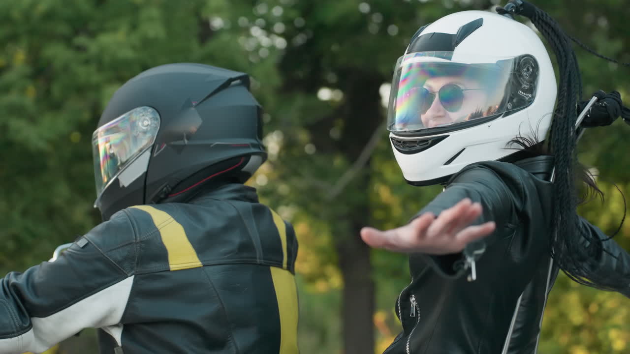 Close up of motorcycle rider in black helmet and leather jacket holding handlebars while passenger behind spreads hand during journey on road surrounded by green trees