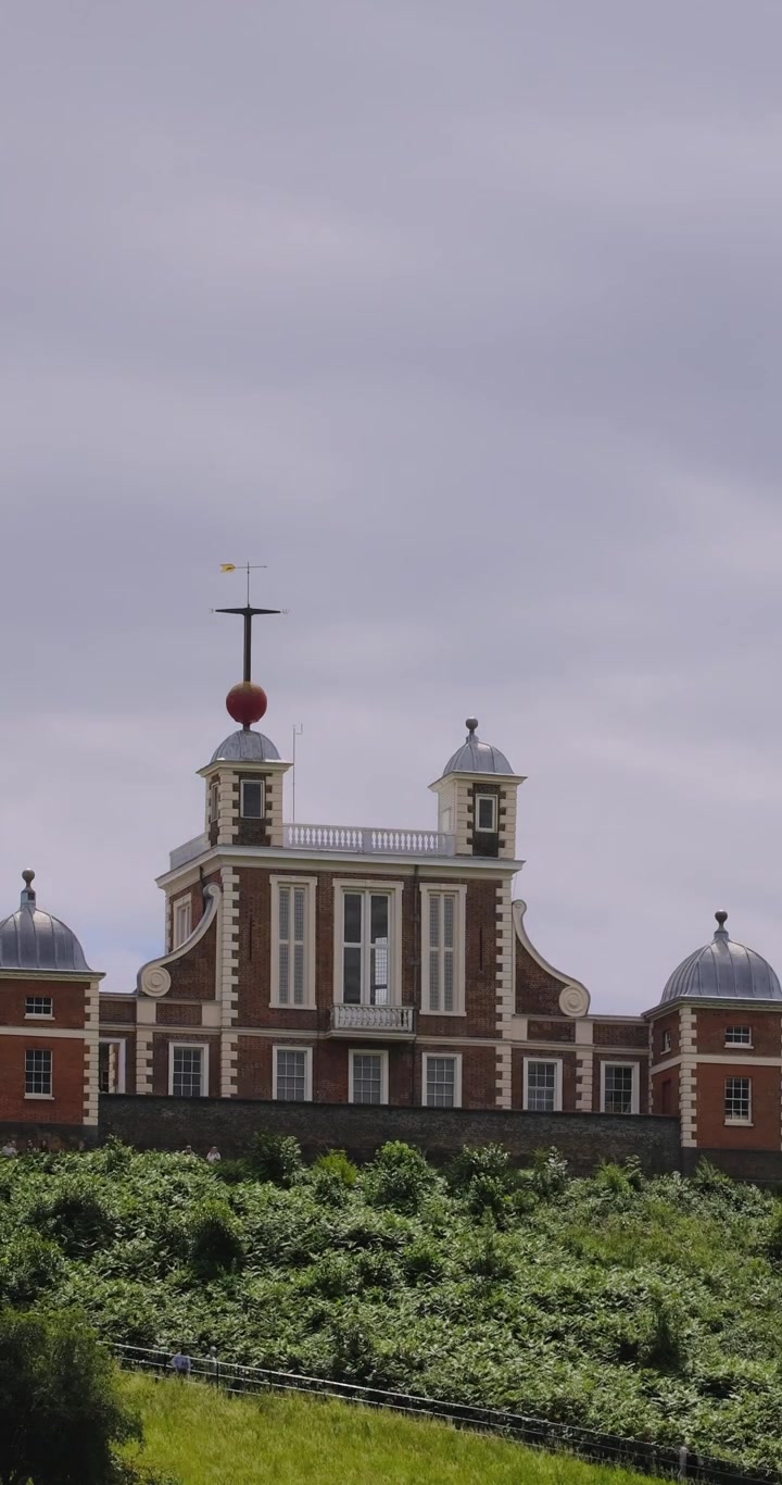 Timelapse vertical da Flamsteed House no Observatório Real de Greenwich com nuvens flutuando acima e visitantes caminhando pelos terrenos históricos.