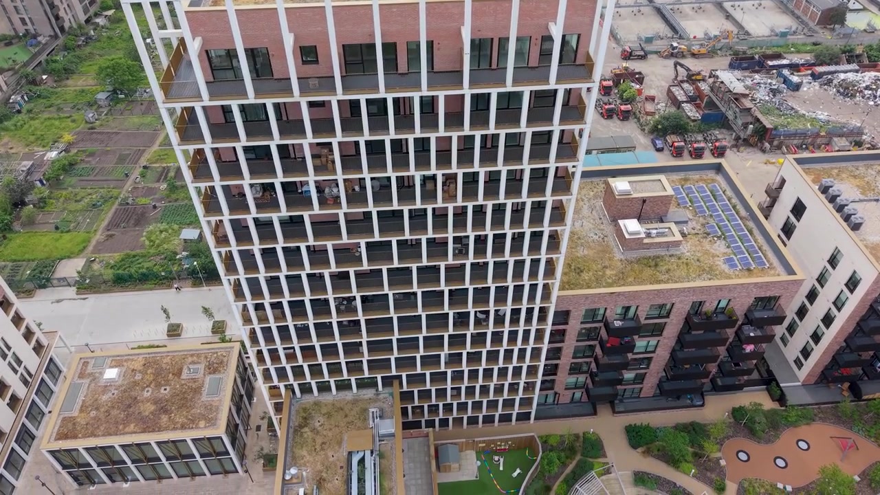 Ripresa con drone in avvicinamento e inclinazione verso il basso del grattacielo Wigeon Heights, da una vista frontale a una dall'alto, che termina sopra la facciata a griglia geometrica e i tetti, evidenziando l'architettura residenziale moderna.