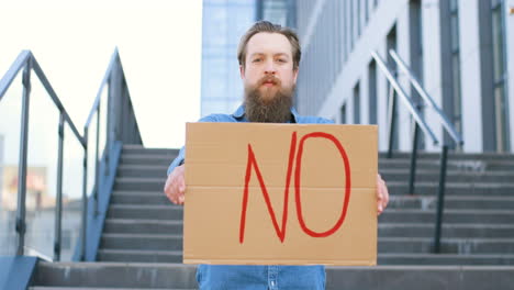 Close-up-view-of-bearded-caucasian-man-holding-No"-signboard-and-looking-at-camera-in-the-street"