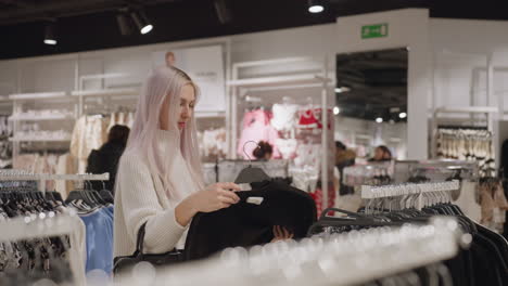 sophisticated shopper holding black polo on hanger returns it to rack in bright boutique, browsing neutral garments under warm spotlights while carrying chic leather handbag and evaluating texture