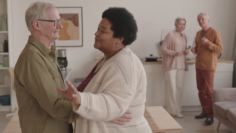 medium shot of joyful african woman slow dancing with her senior caucasian husband at home, couple of their friends smiling, looking at them, standing on background
