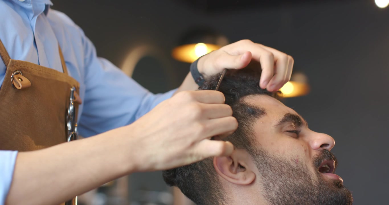 Mandlig barber og kunde i barbersalon, barber løfter krøllet hår, reder og trimmer toppen for at forme.