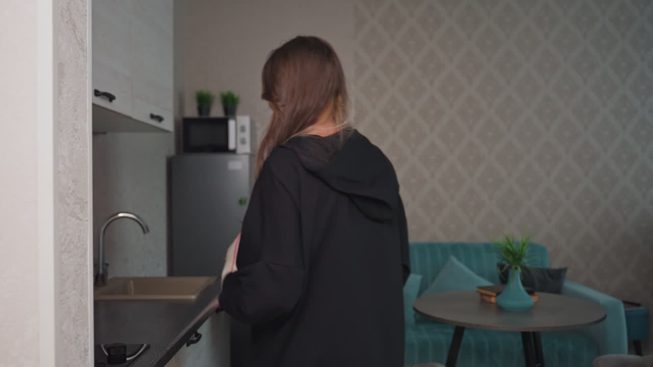 Warm hug between sisters in modern apartment hallway as one places bag and loaf of bread on table and the other fills jug from kitchen tap, capturing cozy moment of reunion and supportive friendship