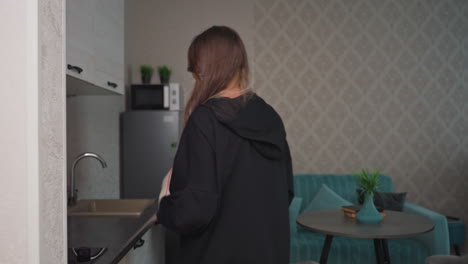 Warm hug between sisters in modern apartment hallway as one places bag and loaf of bread on table and the other fills jug from kitchen tap, capturing cozy moment of reunion and supportive friendship