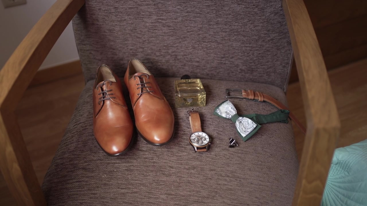 Scarpe da uomo marroni, papillon verde, orologio, cintura, colonia e gemelli disposti ordinatamente su una sedia.
