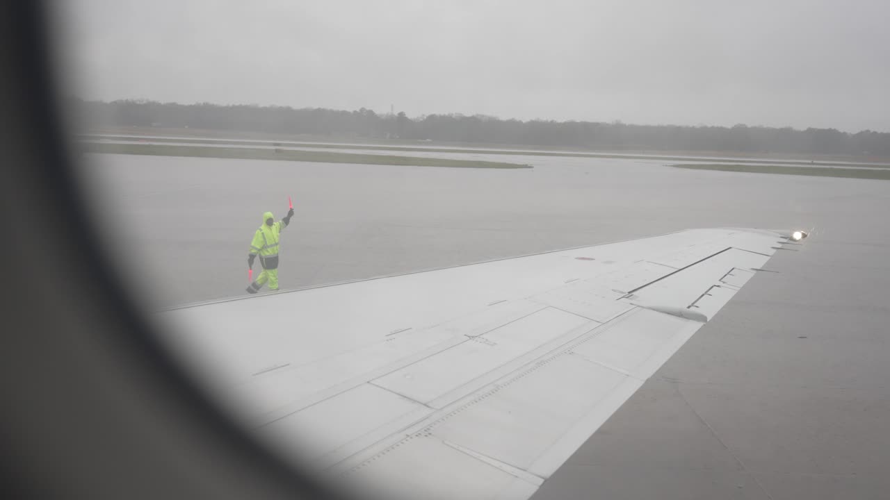 un caminante masculino guía un avión con bastones naranjas en la pista asfaltada antes del despegue, como se ve desde el interior del avión comercial
