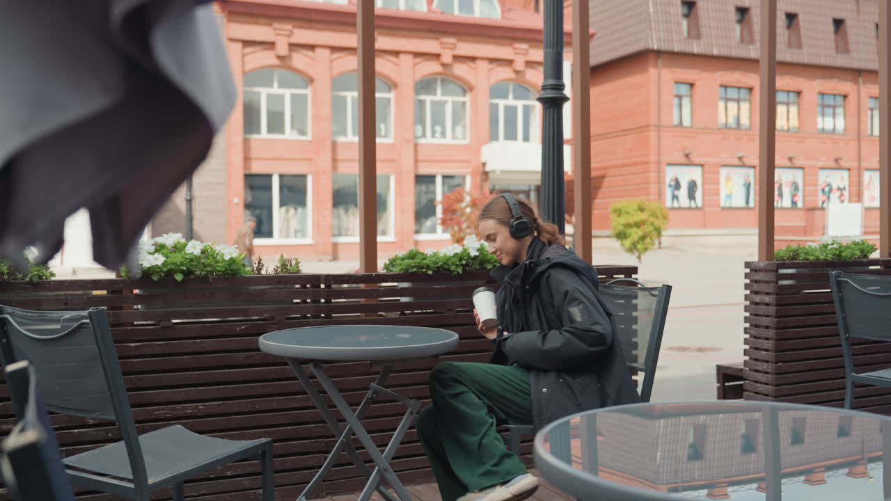 caucasian woman sitting outdoor cafe, arriving then choosing seat, placing bag on chair, unfolding coat, leaning back and sipping coffee from reusable cup, contemplative gaze toward redbrick