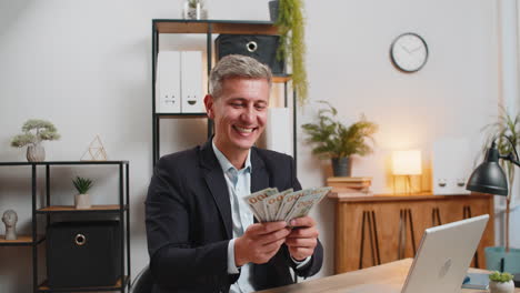 young businessman holding fan of cash money dollar banknotes celebrate success at home office desk