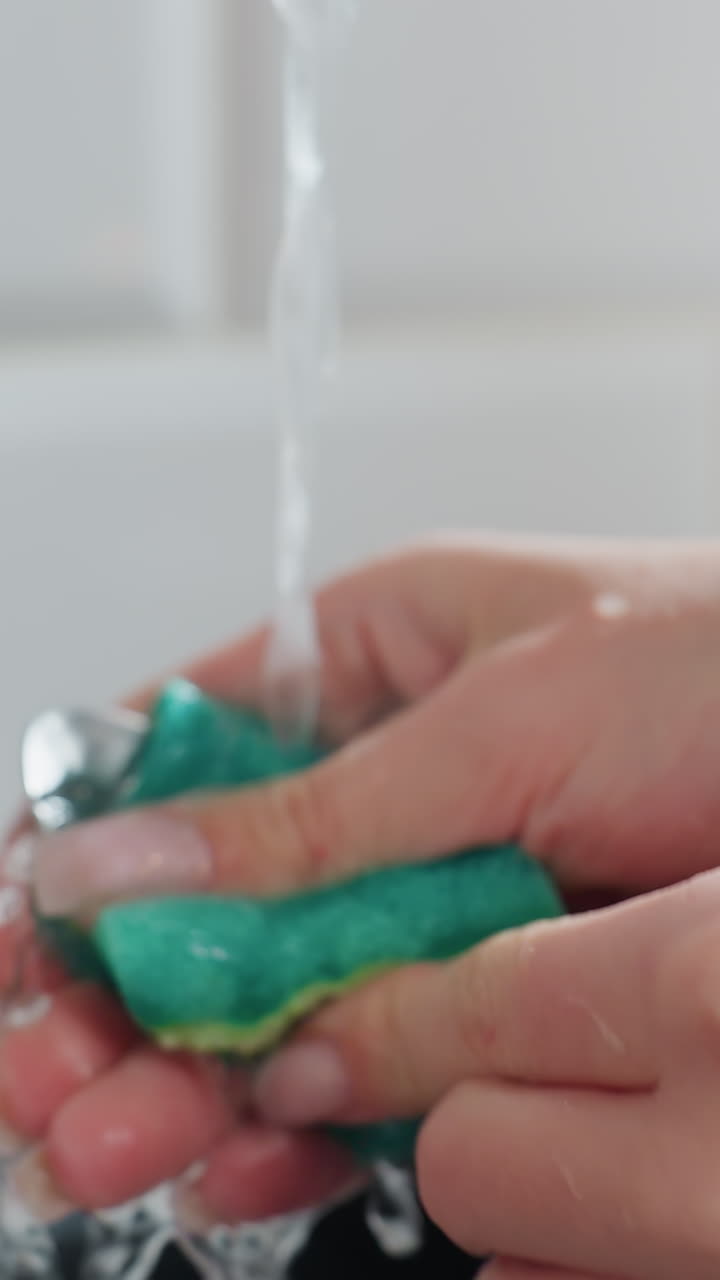 Close-up of hands with a wrist bracelet washing spoon under running tap water, kitchen sink setting, soft focus on hands and water droplets