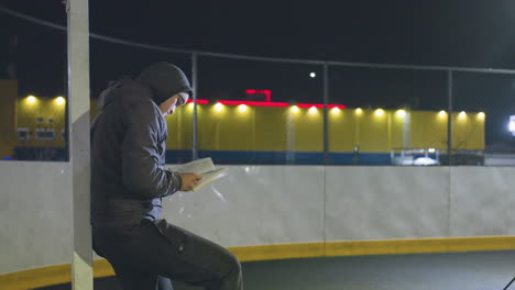 man in hoodie leaning against metallic goalpost outdoors at night reading a book under bright urban lights with a peaceful expression