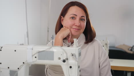 portrait view of seamstress in light top resting hand on sewing machine, intense focused energy visible in eyes, sitting confidently in organized sewing workshop surrounded by tailoring tools